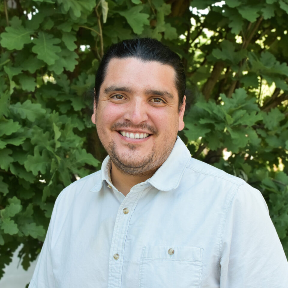 Jose smiling into the camera wearing a white collared shirt and standing in front of greenery.