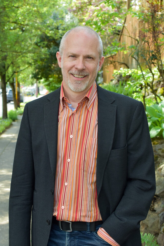 Michael smiling at the camera while wearing an orange button up and black blazer, standing on the sidewalk outside with trees in the background.