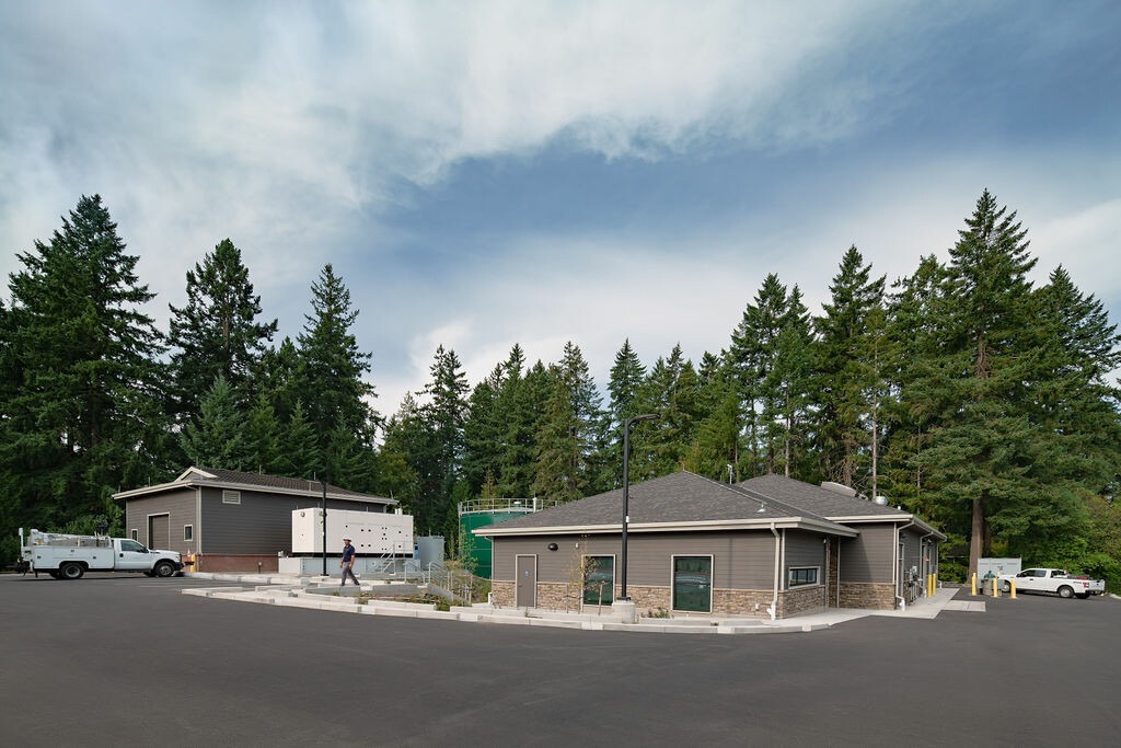 One story building with brick skirting. In the background is a large water tank and evergreen trees.